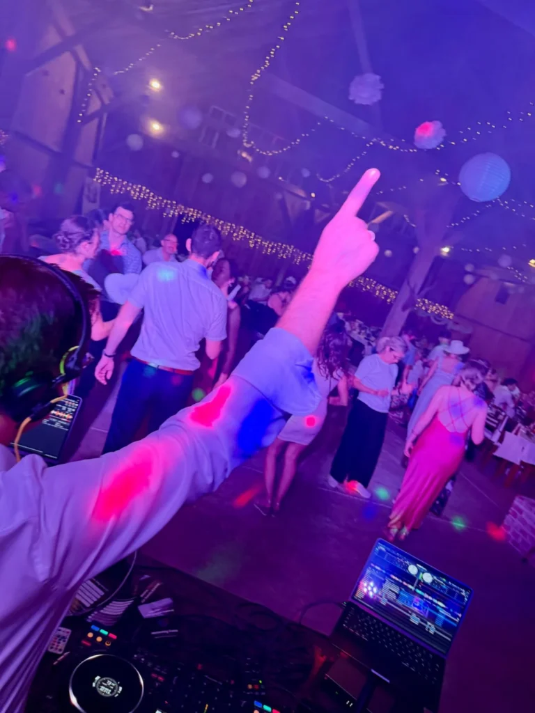 Blick vom DJ-Pult auf die volle Tanzfläche einer Hochzeit in einer Scheune mit Lichterketten